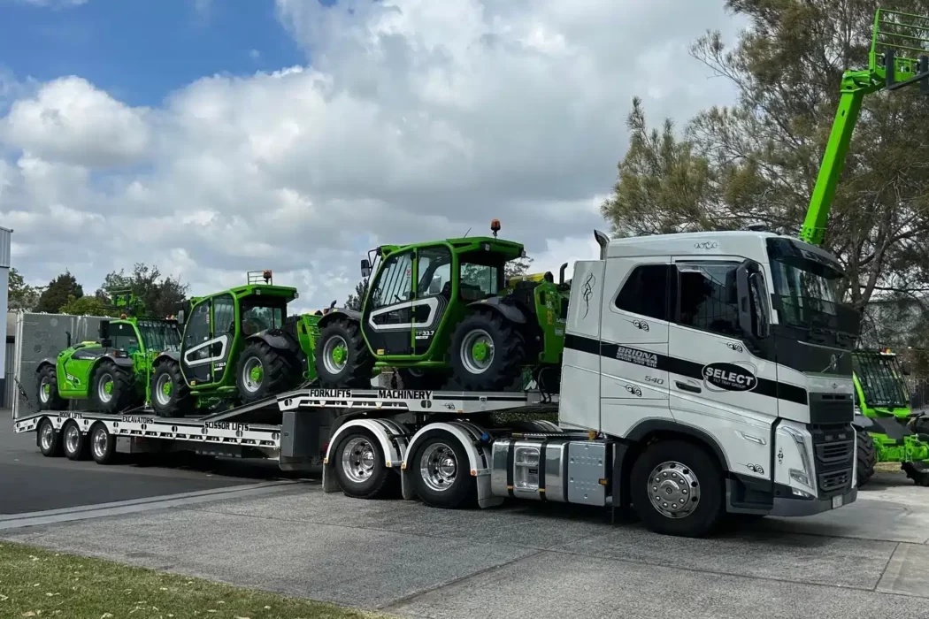 A prime mover towing 3 green heavy machinery on a flatbed trailer